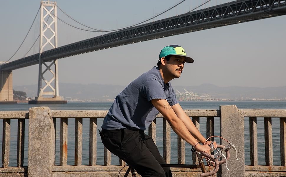 masculine person wearing the Coal Provo hat while riding a bicycle with a bridge in the background